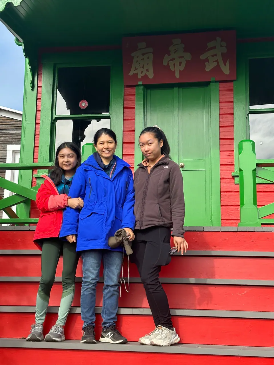 A girl in red, a woman in blue and a girl in a gray fleece stand in front of the green doors of a small red-and-green building.