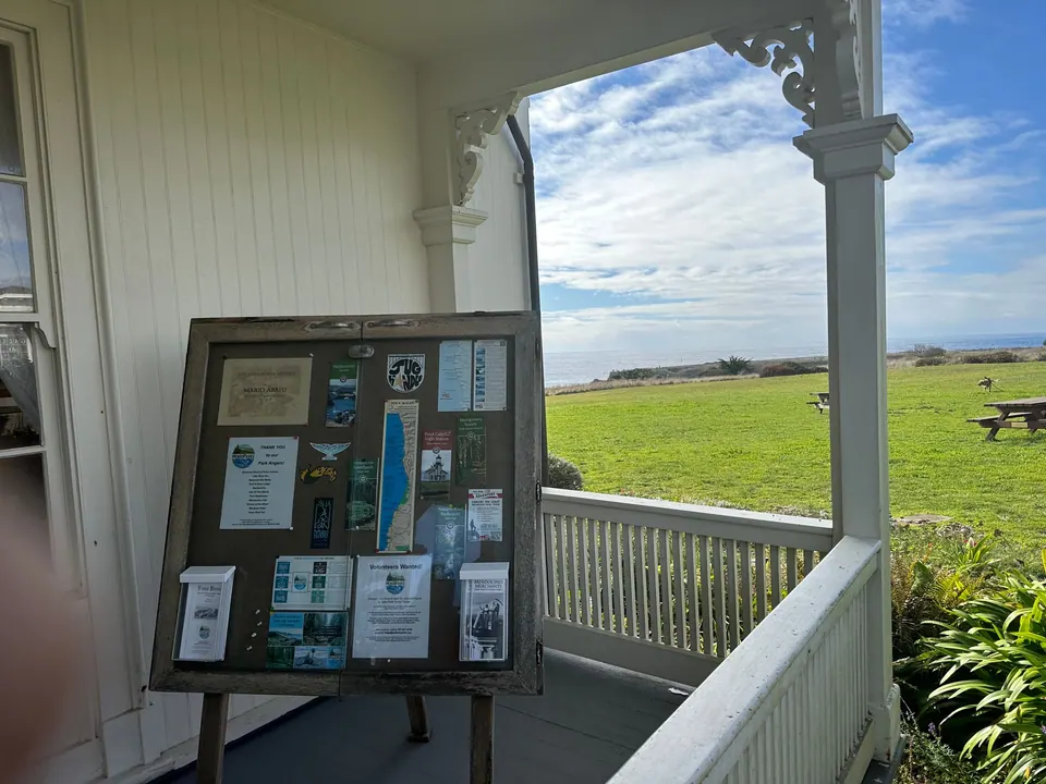 A notice board with maps and leaflets on a white porch, and a lawn with picnic tables running toward the sea.