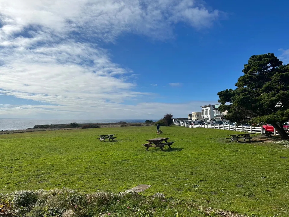A wide green lawn with picnic tables and a white fence, blue sky and clouds above, the ocean on the horizon.