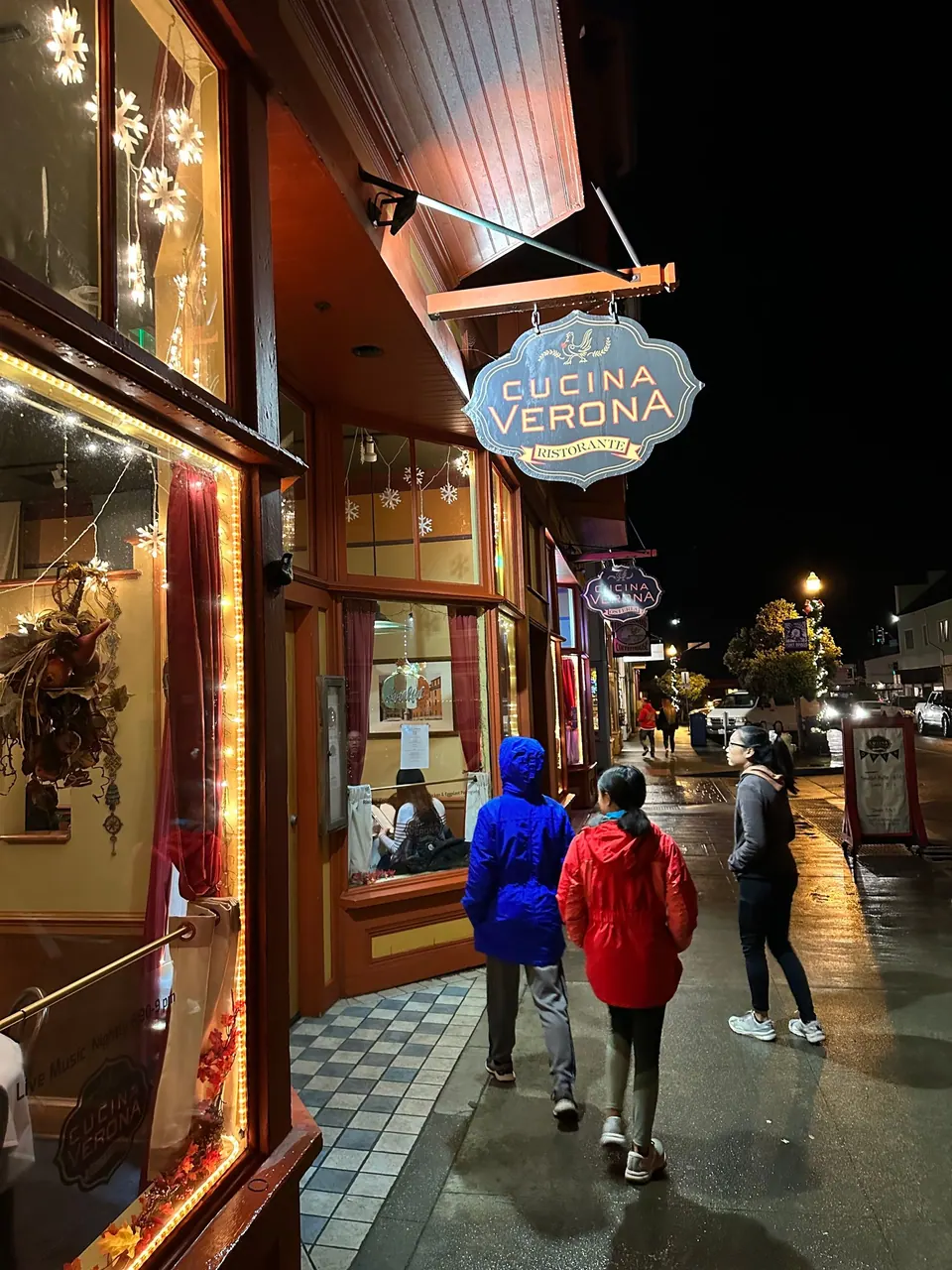 Three people in jackets walk away down a wet sidewalk beneath a hanging sign that reads Cucina Verona.