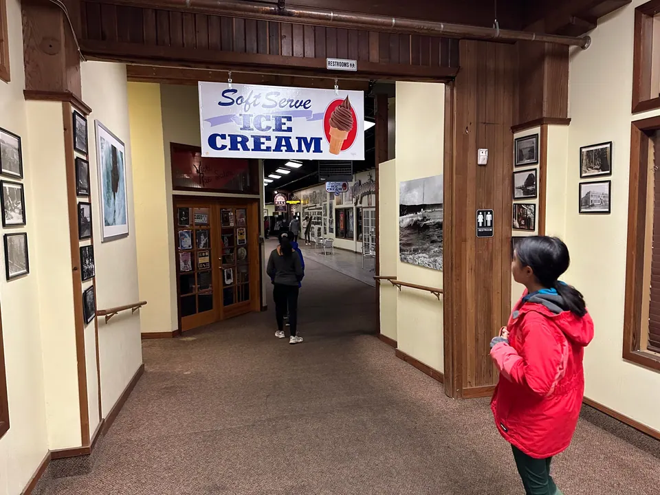 A girl in a red jacket stands in the entrance of a covered arcade of shops with framed photos on the walls, under a sign reading Soft Serve Ice Cream.