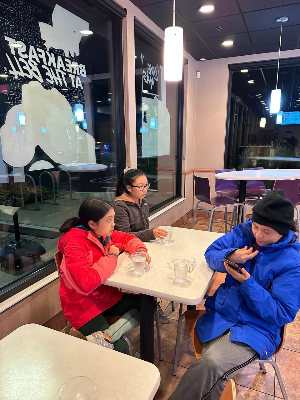 A girl in a red jacket, a girl with glasses and a person in a blue puffer jacket sit at a white table with water glasses, a window sign reading Breakfast at the Bell behind them.
