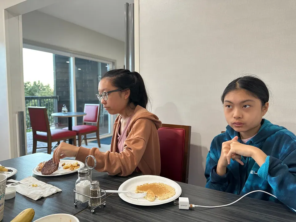 A girl with glasses in a brown hoodie and a girl in a blue tie-dye hoodie sit at a table with a pancake, sausage and a banana.