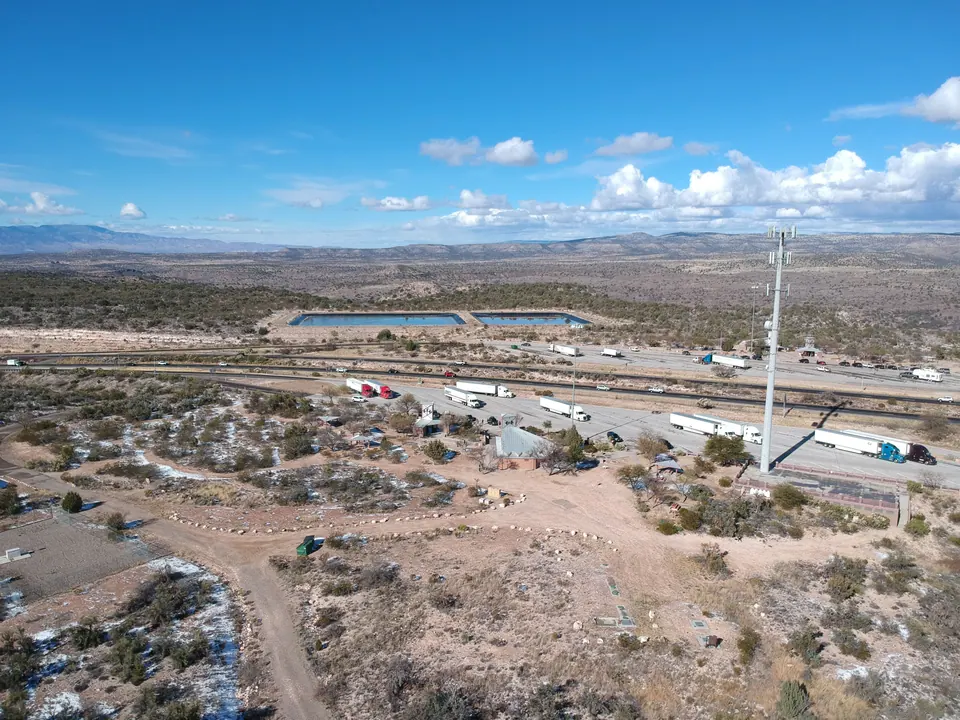 Aerial view of high desert with patches of snow, a dirt yard, a cell tower and big trucks parked along a highway under white clouds.