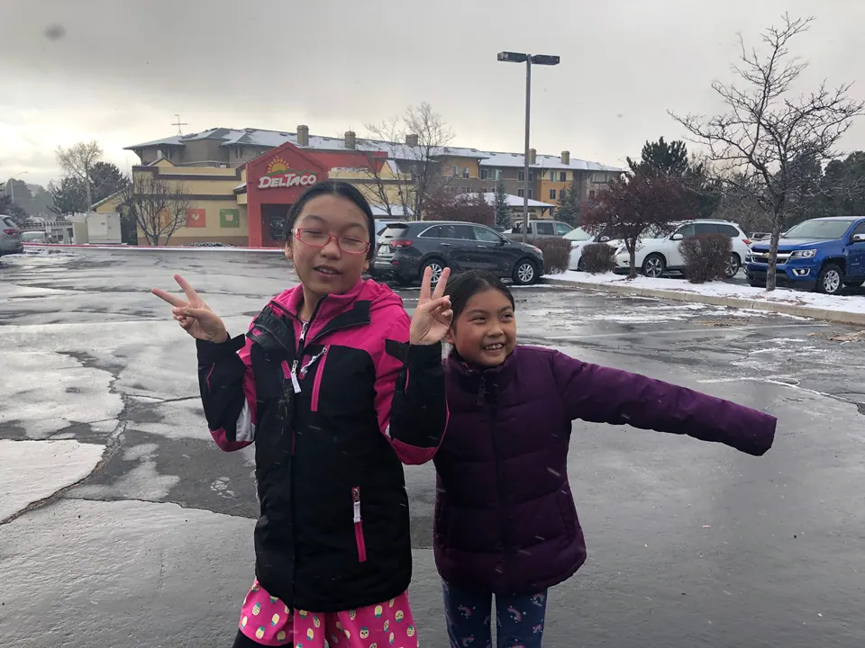 Two girls in winter jackets clown for the camera on wet pavement, one with peace signs and one with her arms out, a Del Taco and snow-dusted cars behind them.