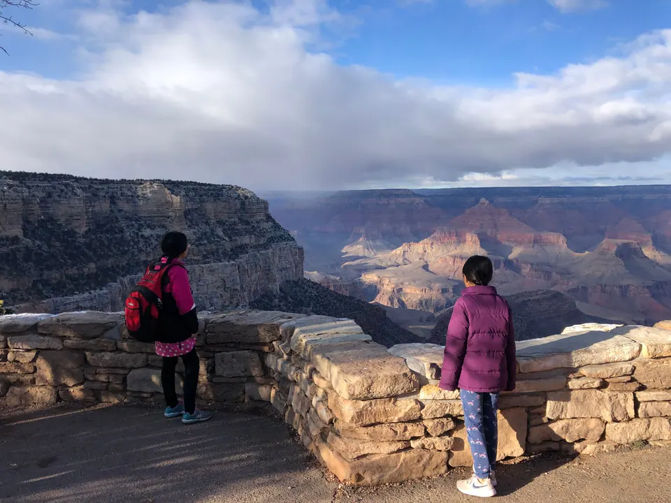 Two girls seen from behind lean on a low stone wall and look out over a vast canyon under a large bank of cloud.
