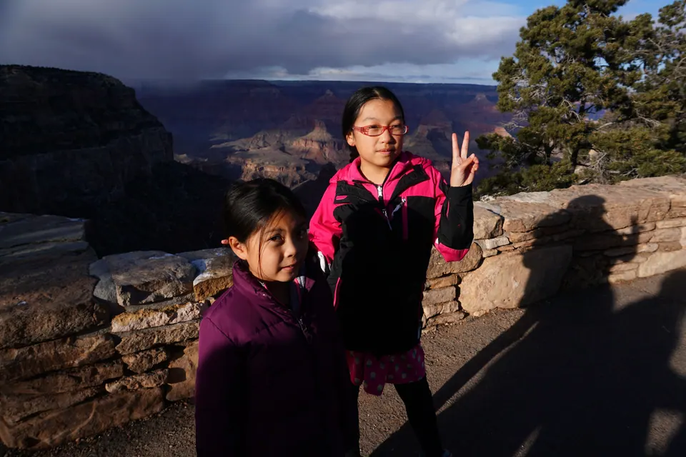 Two girls in purple and pink jackets pose by a stone wall, one with a peace sign, with dark cloud over the canyon's buttes behind them.