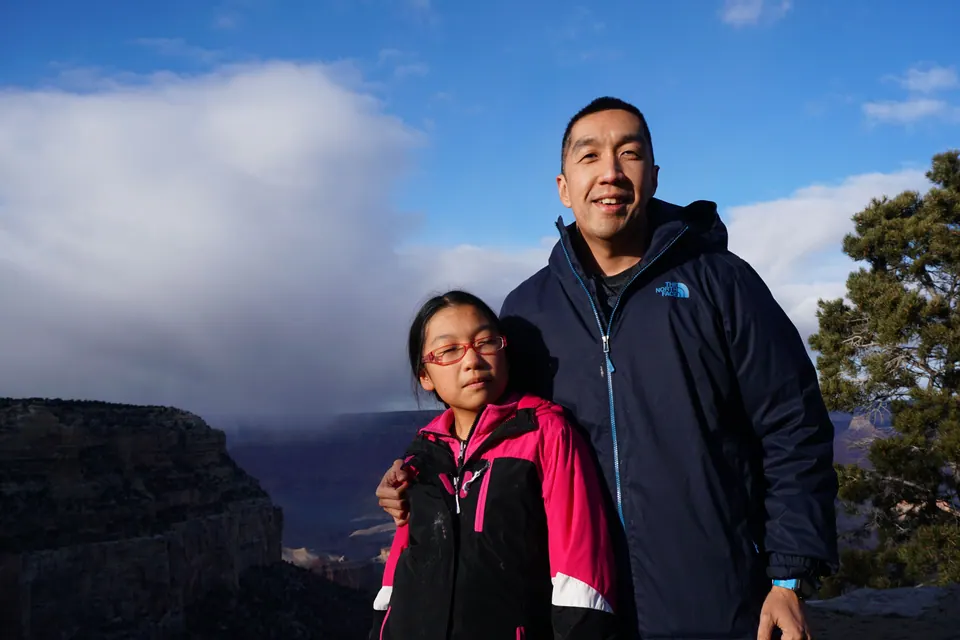 A smiling man in a navy jacket with his arm around a girl in glasses and a pink jacket, a towering white cloud over the canyon behind them.