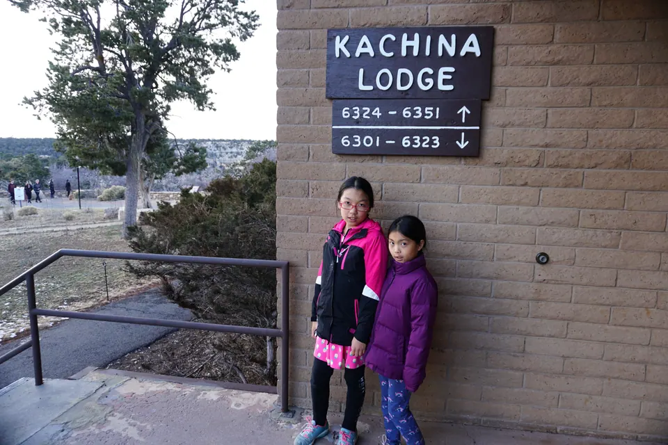 Two girls stand against a tan brick wall beneath a wooden sign reading Kachina Lodge with room numbers and arrows.