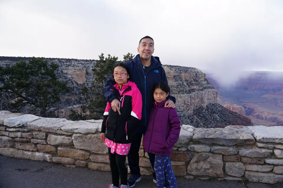 A man and two girls stand at a low stone wall, with cliffs, pines and mist filling the canyon behind them.