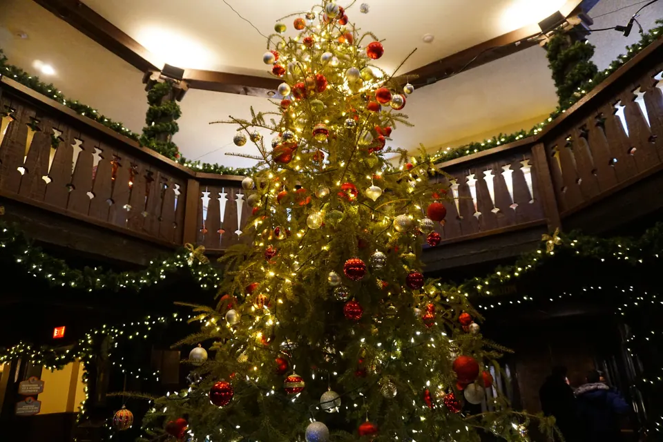 A tall Christmas tree with red and gold ornaments and white lights rises past a dark wooden balcony with cut-out railings hung with garlands.
