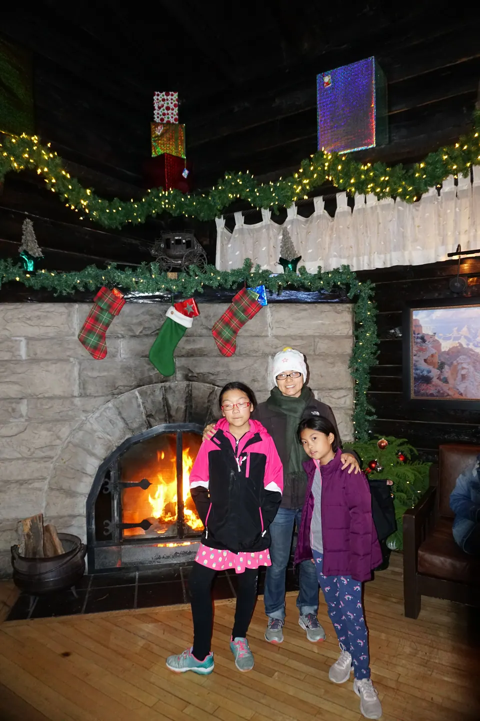 A woman in a white knit hat and two girls stand in front of a stone fireplace with a fire burning and Christmas stockings on the mantel.