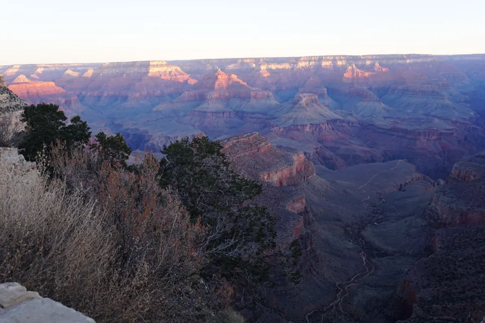 Early sun lights the tops of the canyon's buttes while the depths are still in blue shadow; a trail winds down a slope below the rim.