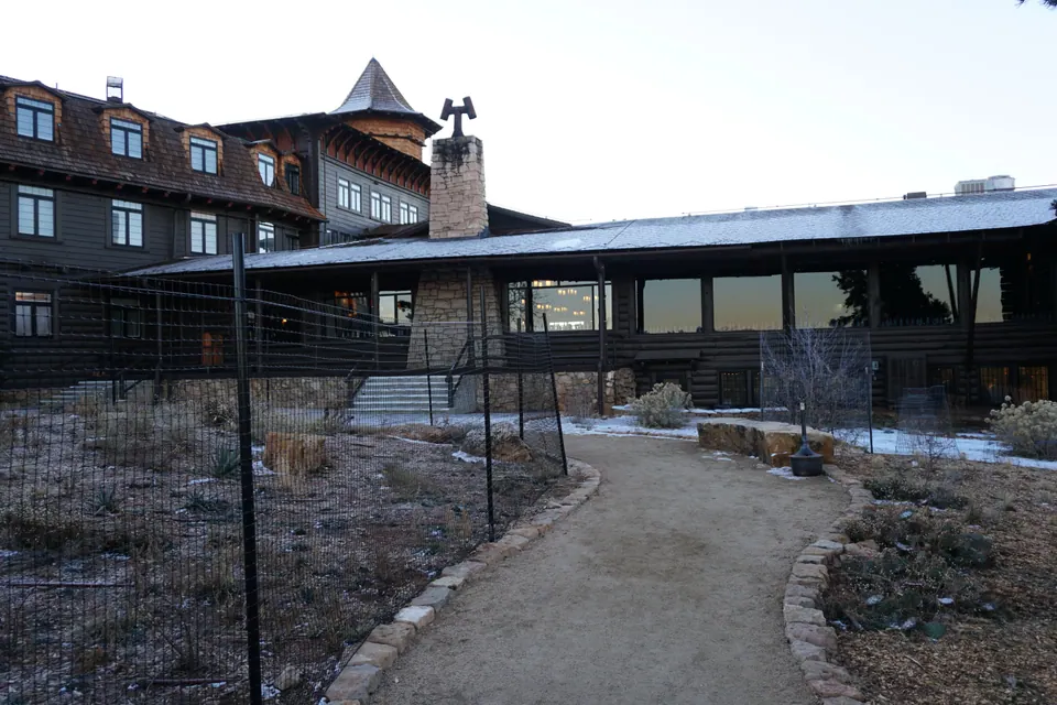 A dark shingled hotel with dormers, a turret and a long log porch at first light, with frost and a little snow along a garden path.