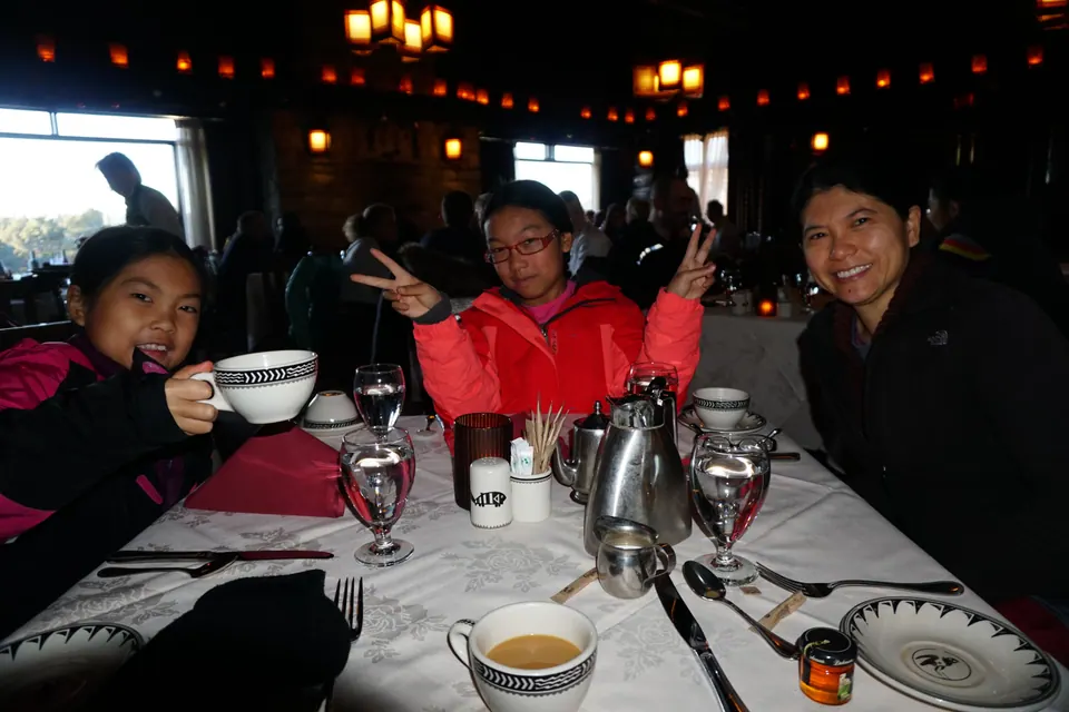 A woman and two girls at a white-clothed table under hanging lanterns, one girl raising a patterned coffee cup and the other making peace signs.