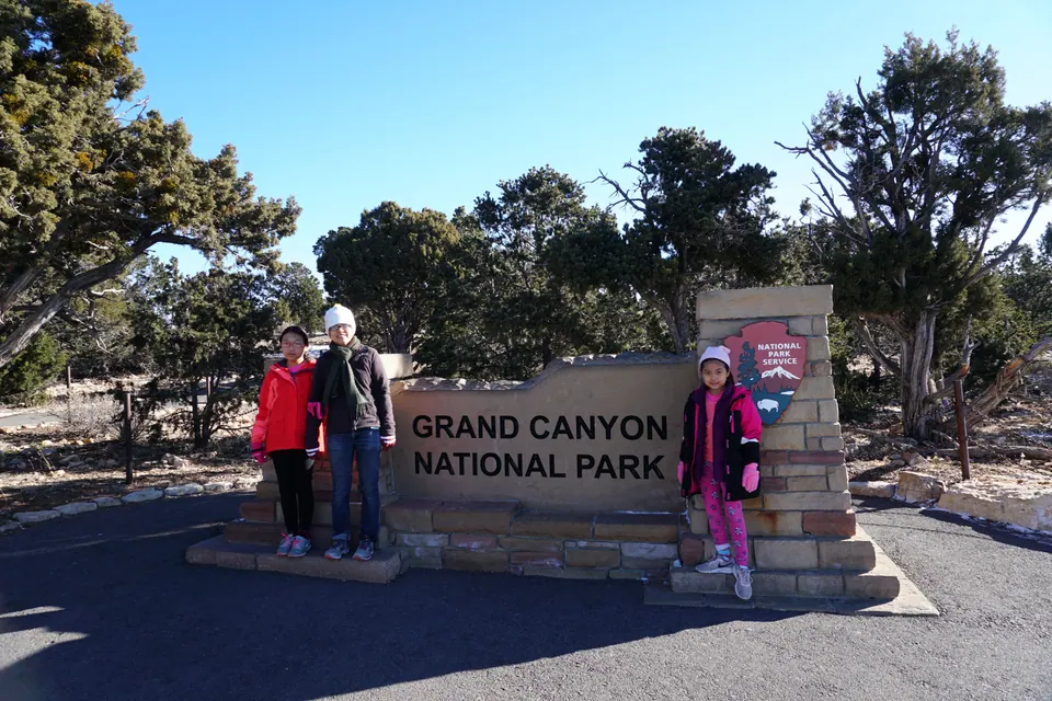 A woman and a girl stand at one end of a stone sign reading Grand Canyon National Park and another girl beside the Park Service arrowhead, under a clear blue sky.