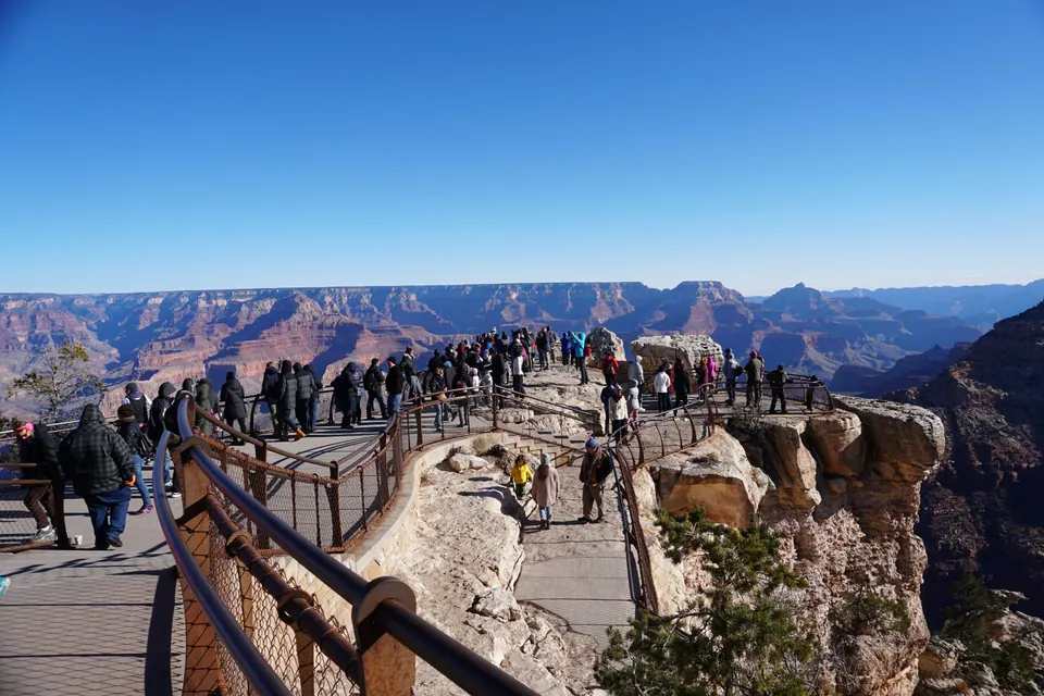 People line railed walkways out onto a rocky promontory, with the canyon spread out under a cloudless sky.