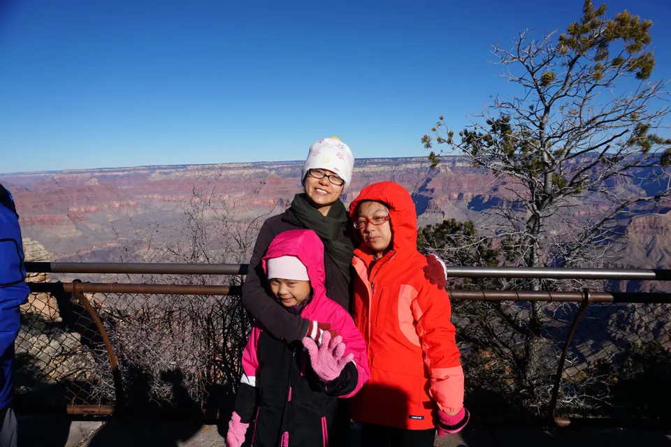 A woman in a white knit hat and two girls in bright pink and orange coats pose at a railing, the canyon behind them in clear morning light.