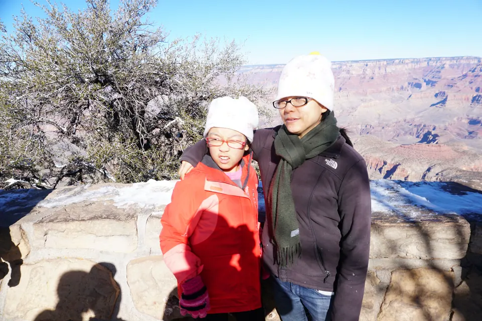 A woman in a white hat and green scarf with her arm around a girl in a red coat at a snowy stone wall, the canyon behind them.