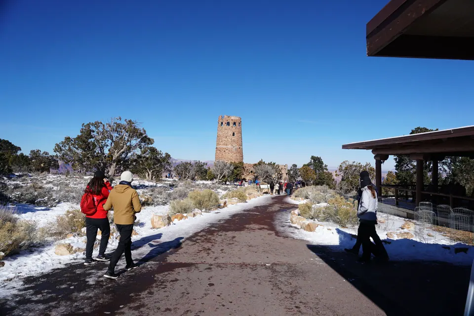 A paved path between snowy banks leads toward a round stone tower under a deep blue sky, with people walking along it.