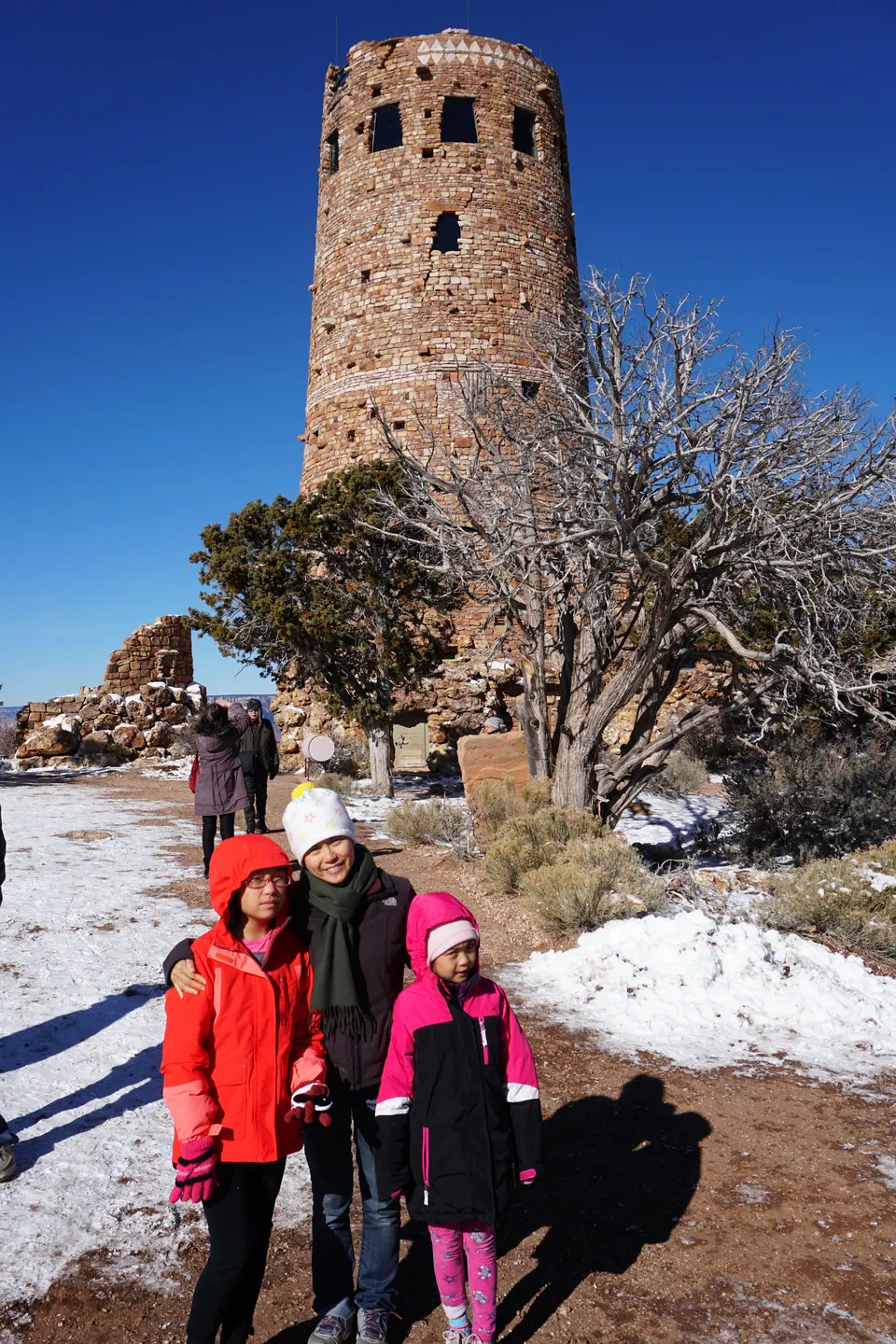 A woman and two girls in bright winter coats stand in front of a tall round tower of stacked stone, with a twisted bare juniper and snow on the ground.