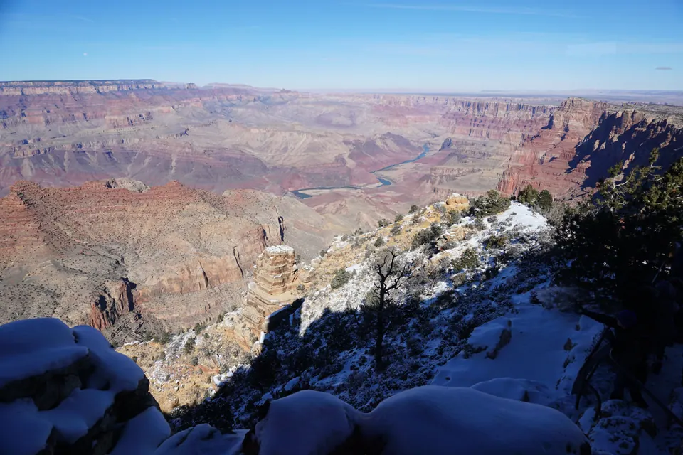 A deep, wide canyon of red and cream cliffs under a clear blue sky, with a river winding along the bottom and snow on the rocks in the foreground.