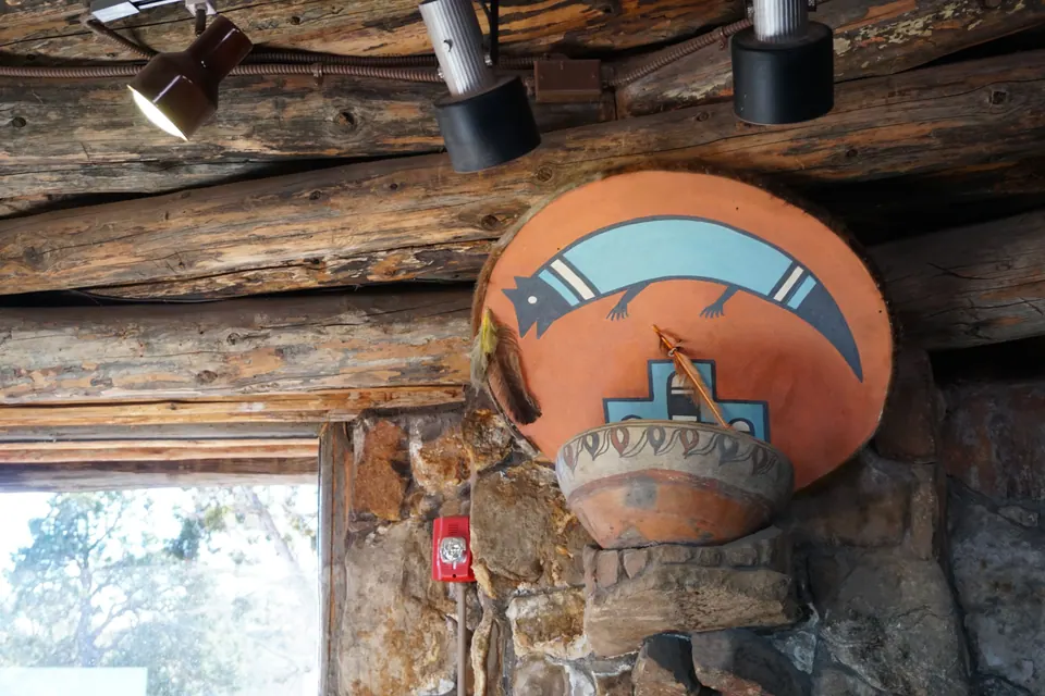 A painted orange disc with a turquoise lizard hangs above a clay bowl on a rough stone wall under log beams.