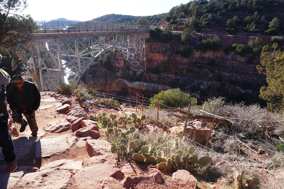 A steel arch bridge spans a red-rock canyon, seen from a rocky path with prickly pear cactus in the foreground.