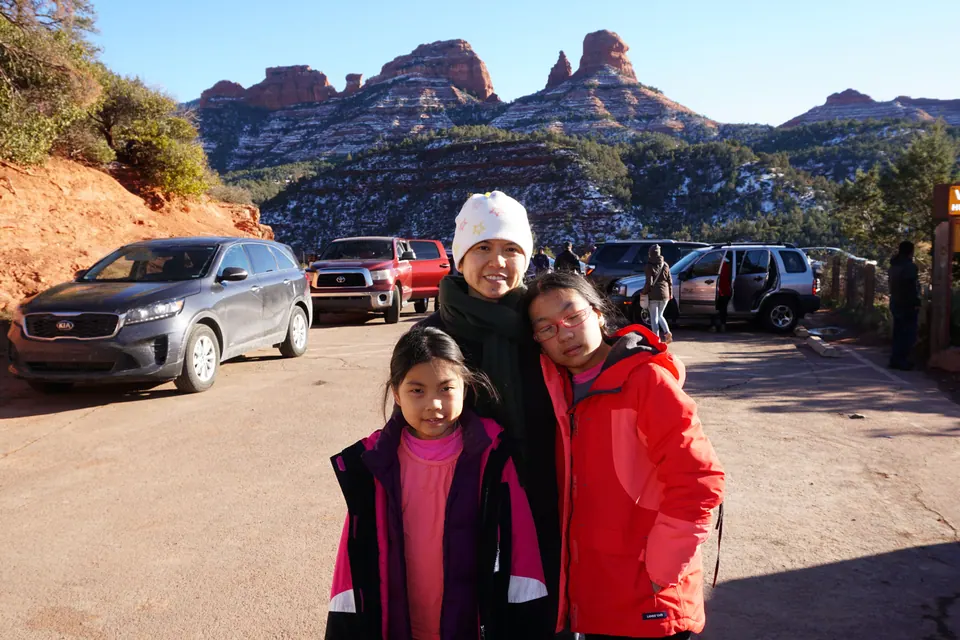 A woman and two girls stand in a dirt parking lot with parked cars, below red-rock spires streaked with snow.