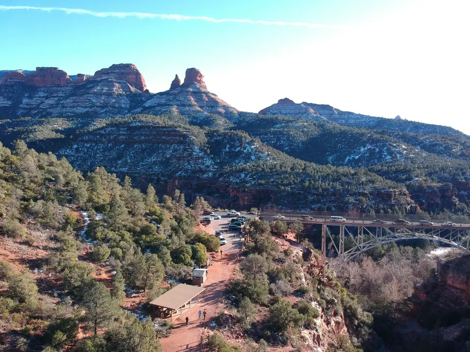 Aerial view of a red-dirt parking area among pines beside a steel arch bridge, with snowy red-rock ridges and spires above.