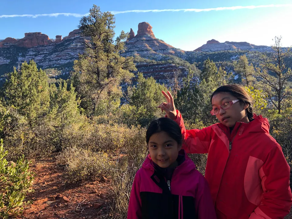 Two girls in pink and red coats, one giving the other bunny ears, with pines, scrub and snow-streaked red rock behind them.