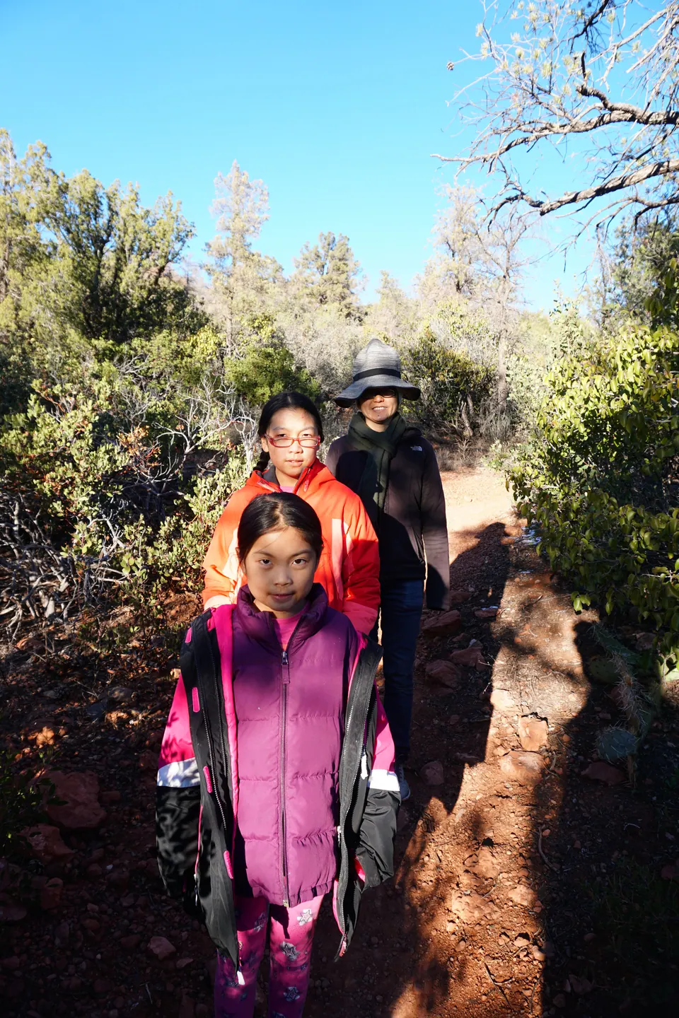 Two girls and a woman in a wide-brimmed hat walk in single file along a narrow red-dirt trail through scrub and pines.