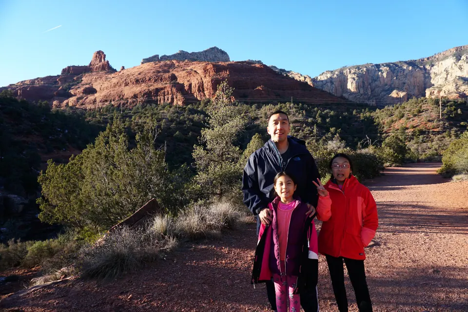 A man and two girls on a wide red-dirt path, a red-rock ridge and pale cliffs behind them in late sun.