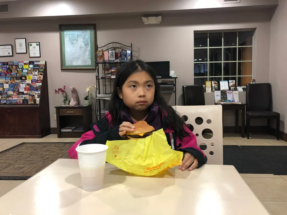 A girl eats a cheeseburger from a yellow wrapper at a table in a hotel lobby with brochure racks and a framed map on the wall.