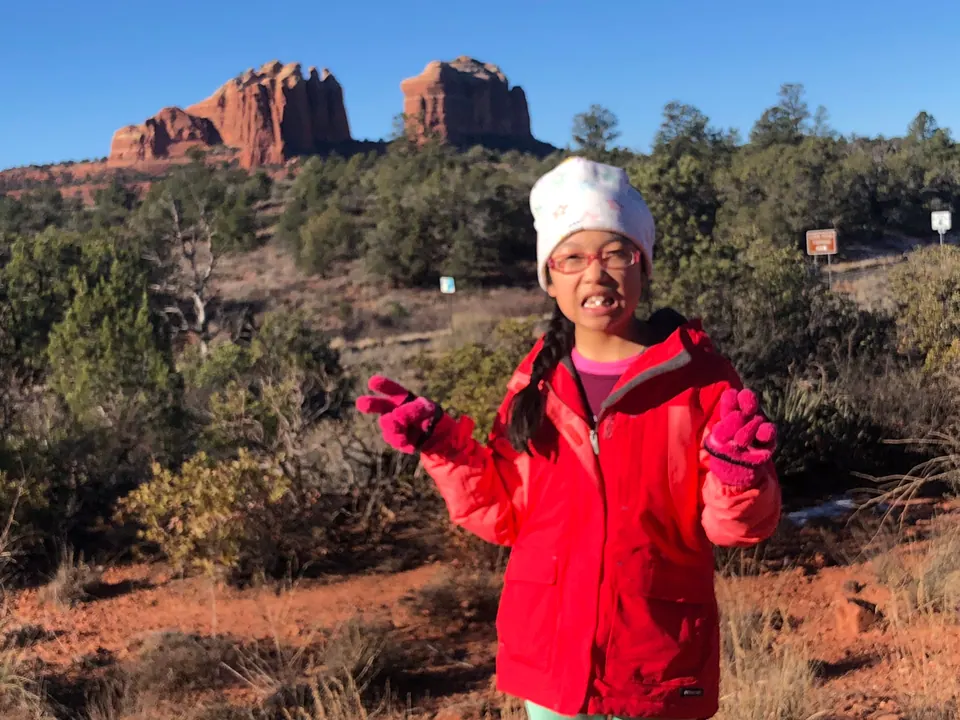A grinning girl in a red coat, pink gloves and a white star-print hat stands among scrub, with two red-rock buttes and trail signs behind her.