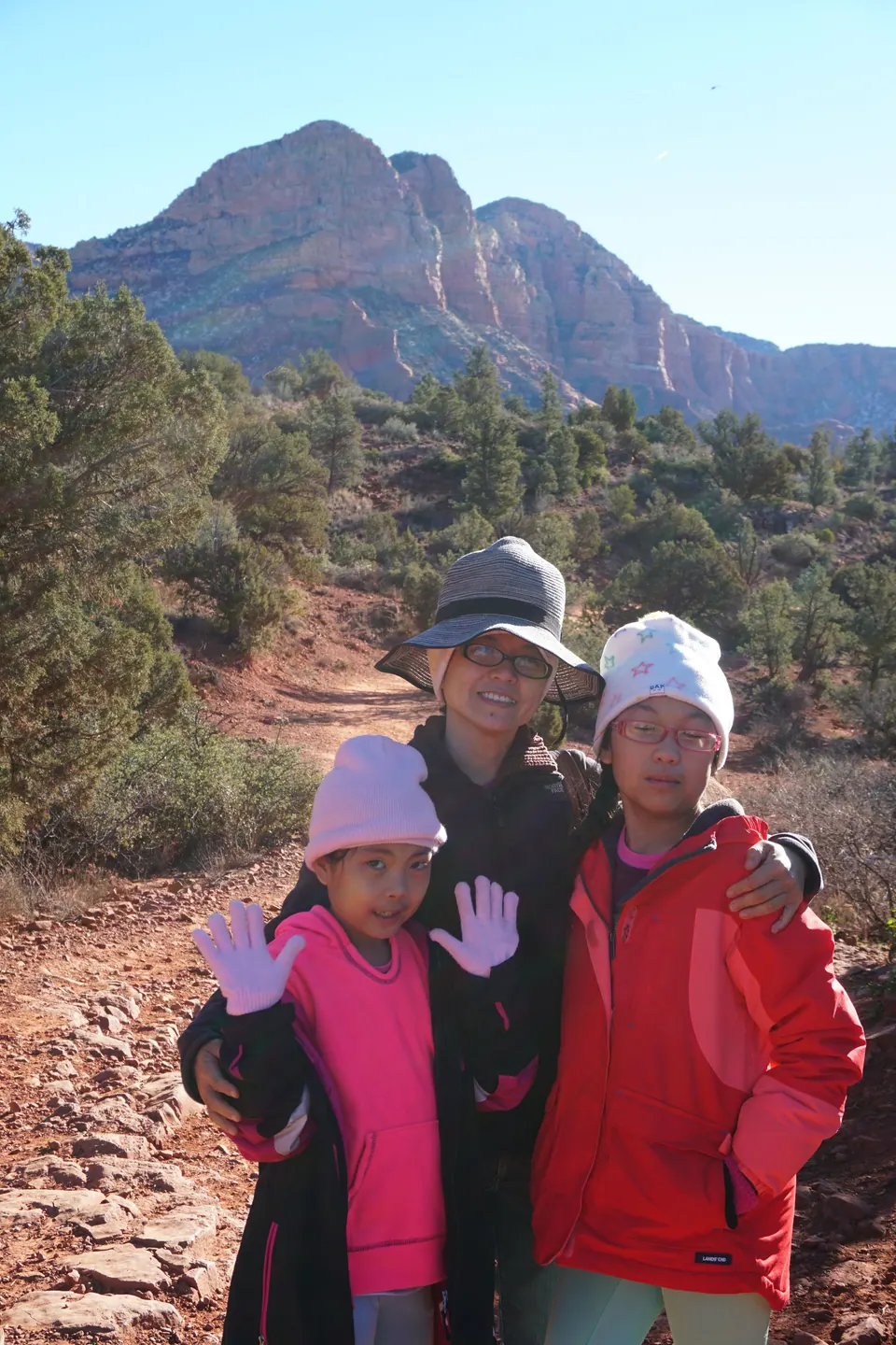 A woman in a striped sun hat with her arms around two girls in winter hats on a rocky red trail, a big red-rock mountain behind them.