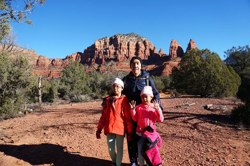 A man and two girls in hats on a wide stretch of red gravel, with a flat-topped red butte and twin spires behind.