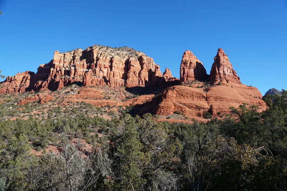 Sunlit red-rock cliffs and two tall pointed spires rise above juniper and scrub under a deep blue sky.