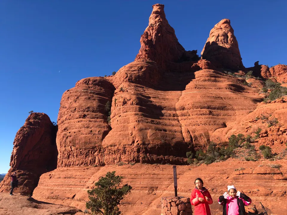 Two girls stand next to a stone cairn with a trail post at the foot of a pair of layered red-rock spires.