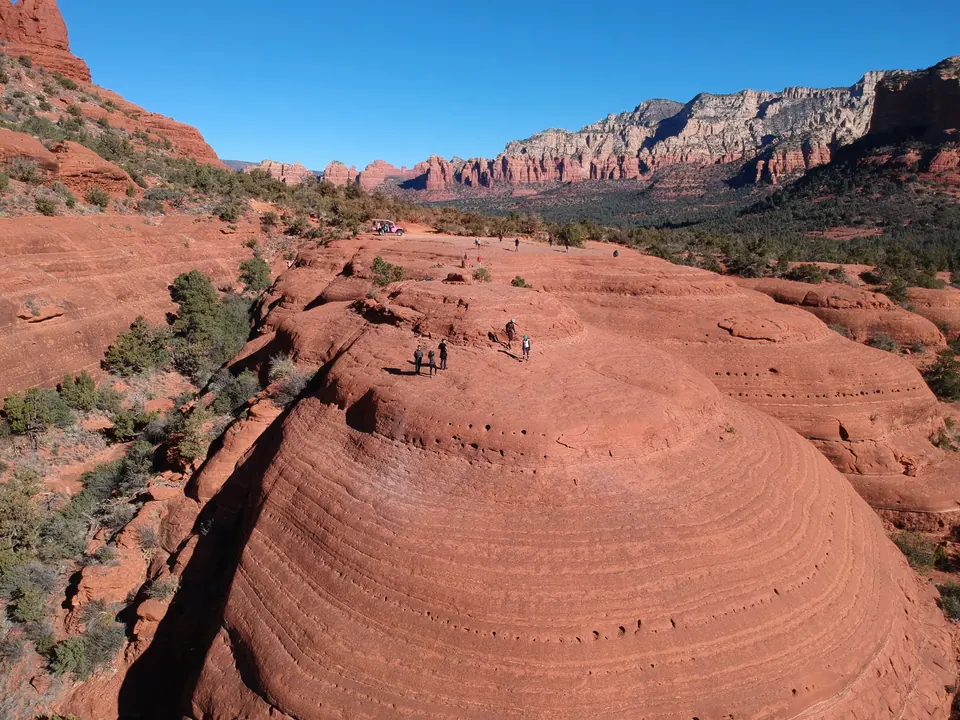 Aerial view over a round, banded red slickrock dome with a few hikers on top, a pink jeep parked beyond, and red and white cliffs across the valley.