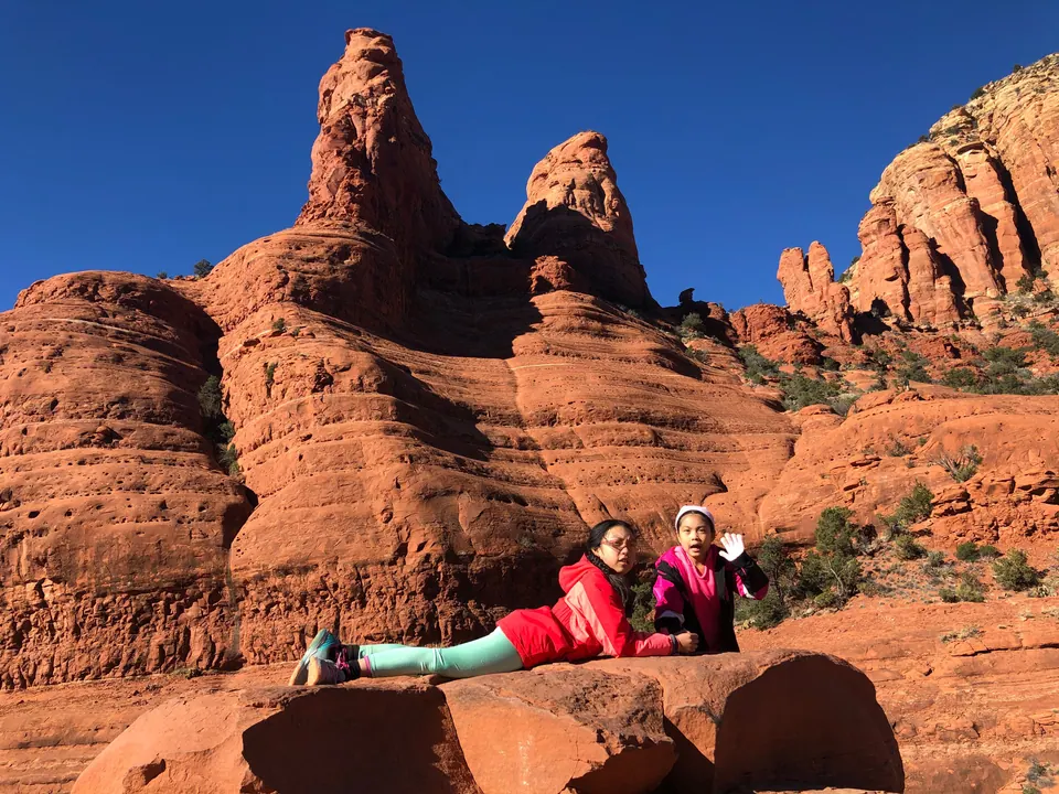 A girl in a red jacket lies on a big red boulder while another girl waves beside her, below two tall layered spires.