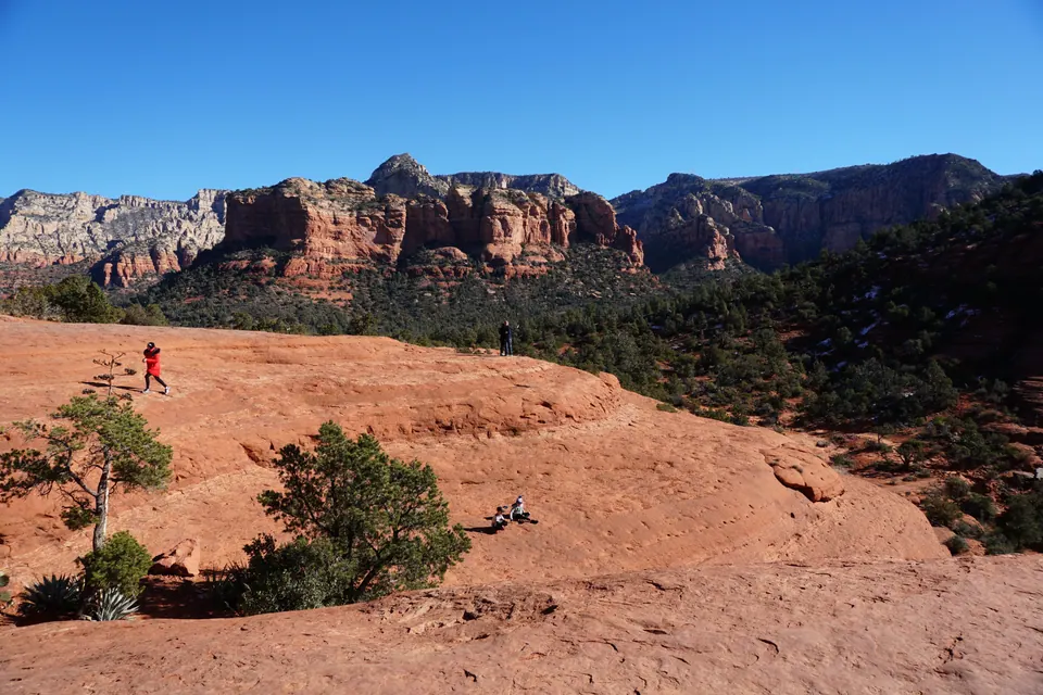 A wide red slickrock slope with a person in red climbing it and two others sitting on the rock, cliffs and forest beyond.
