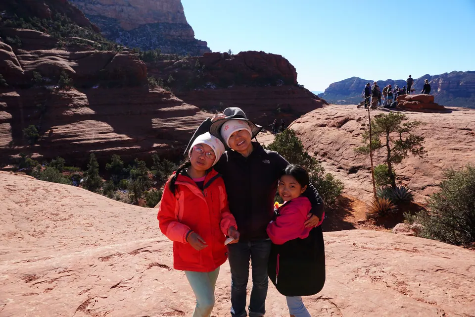 A woman and two girls hug on smooth red rock, with other hikers gathered on a rock ledge behind them.