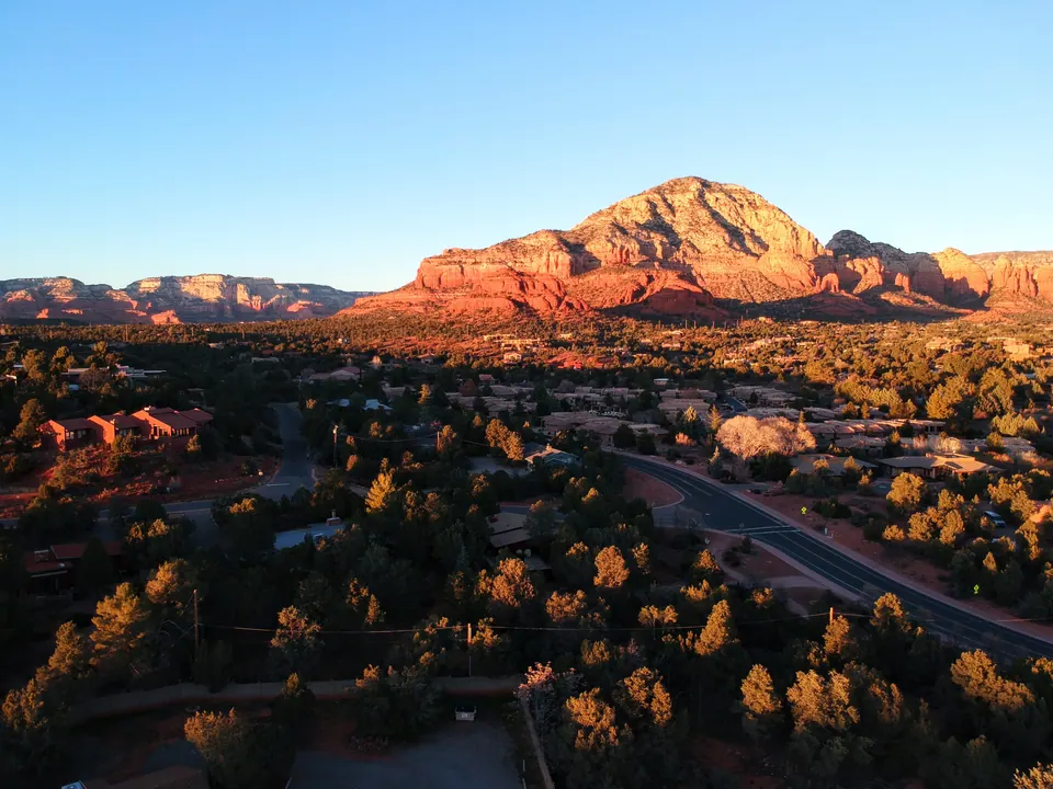 Aerial view at sunset of a red-rock mountain glowing orange above trees, houses and a curving road.