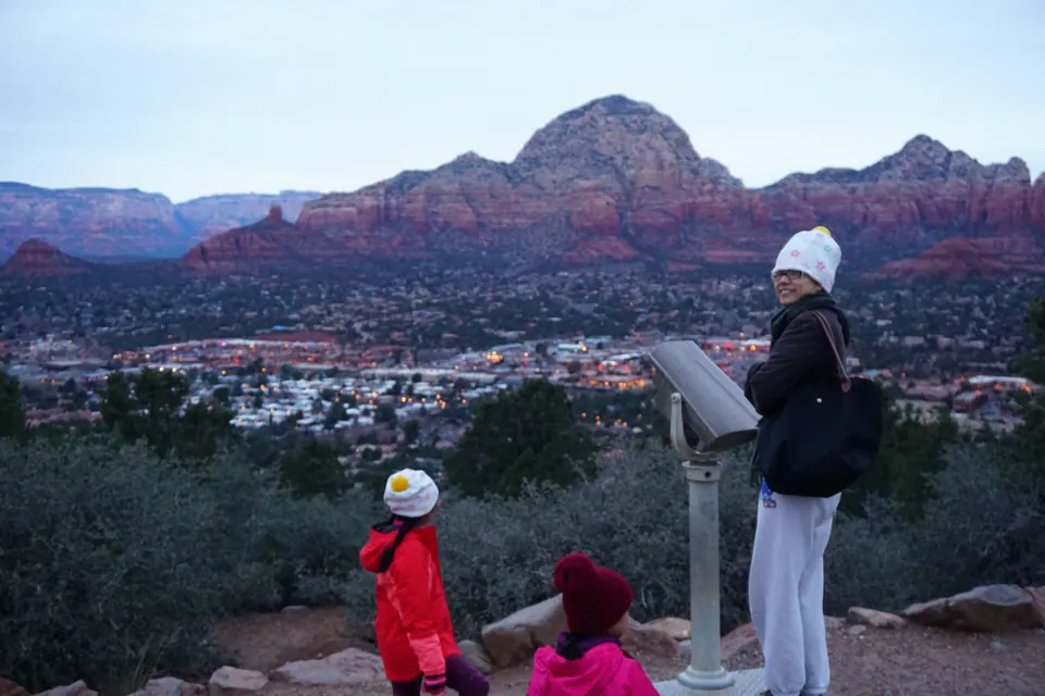 A woman in a white knit hat stands by a coin-operated viewer while two girls look out over the lights of a town below a big red-rock mountain at dawn.