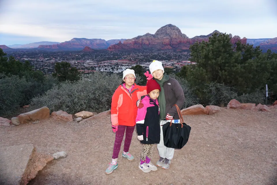 A woman and two girls in winter hats stand on a gravel overlook, with the town and red-rock ridges behind them under grey cloud.