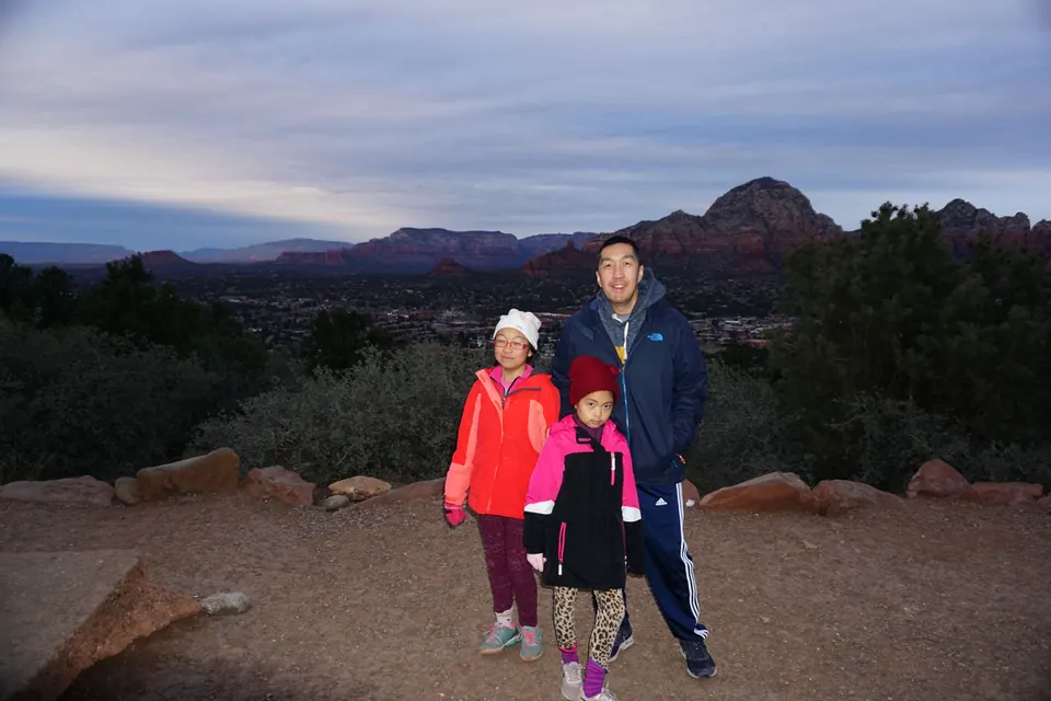 A man in a navy jacket and track pants stands with two girls on a gravel overlook in early morning light, red-rock ridges behind them.