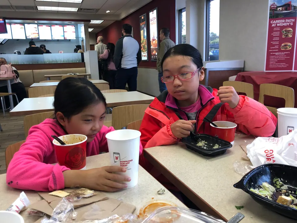 Two girls eat at a fast-food table, one with a red chili cup and a big drink, the other with a salad, Wendy's posters on the wall.