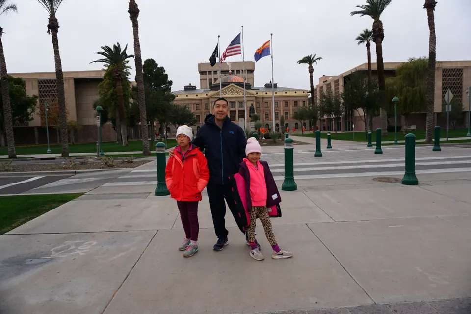 A man and two girls stand on a plaza before a stone capitol building with a copper dome, U.S. and Arizona flags and tall palms.