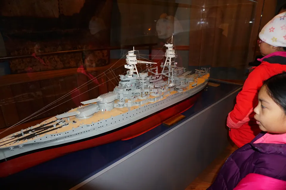 Two girls look at a large model of a gray battleship in a glass case.